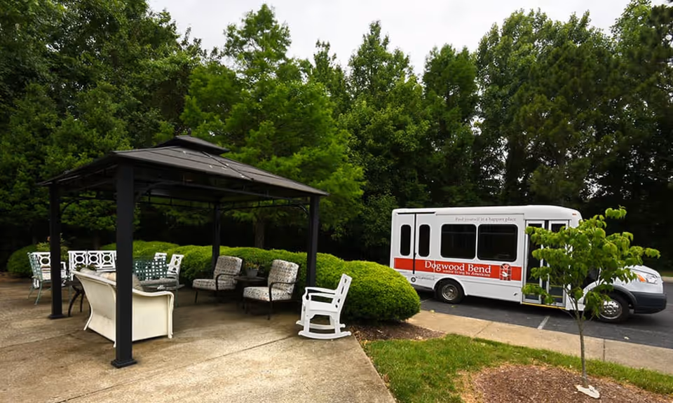 Outdoor seating area with a black metal gazebo covering several chairs and a table, surrounded by green bushes and trees. A white shuttle bus with red accents and the name 'Dogwood Bend' is parked on the adjacent paved area.