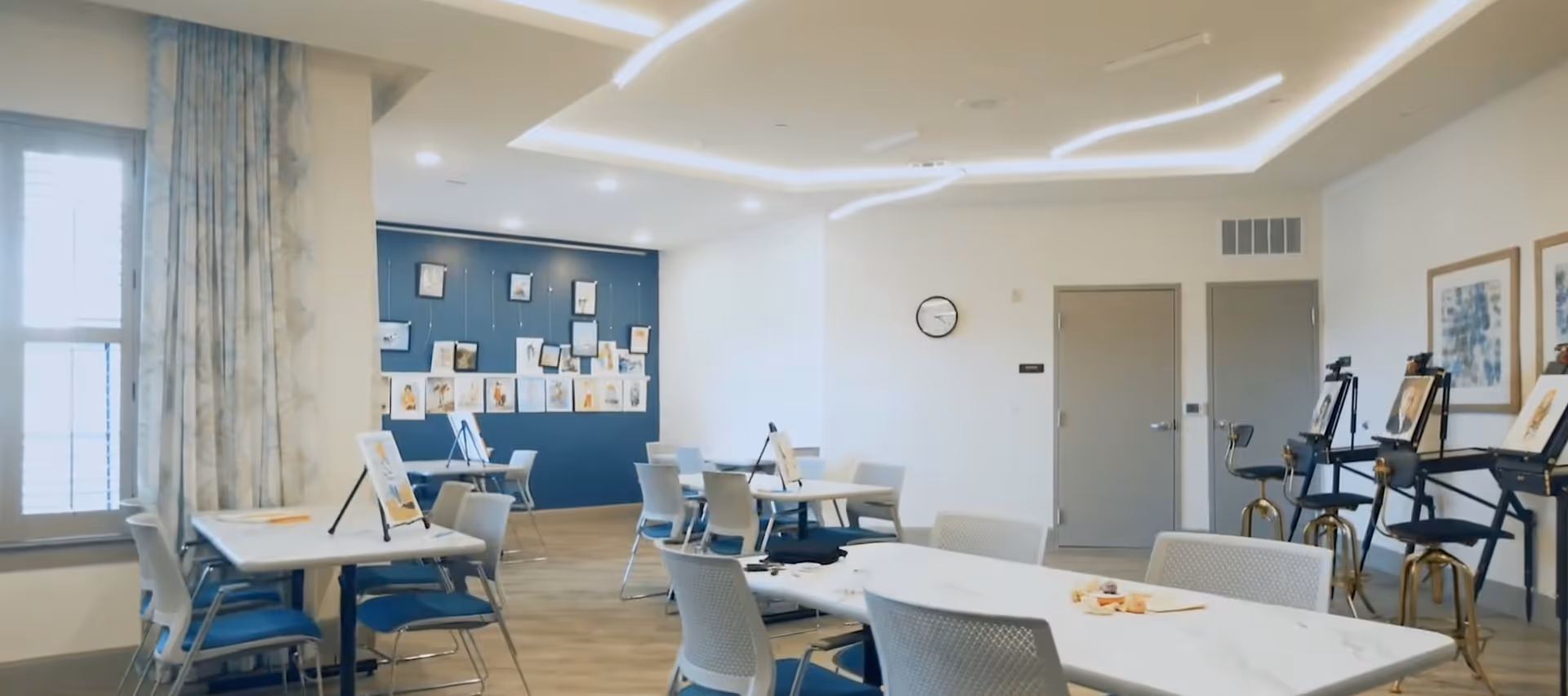 Bright activity/dining room with tables and chairs, art easels, and artwork displayed on a blue accent wall.