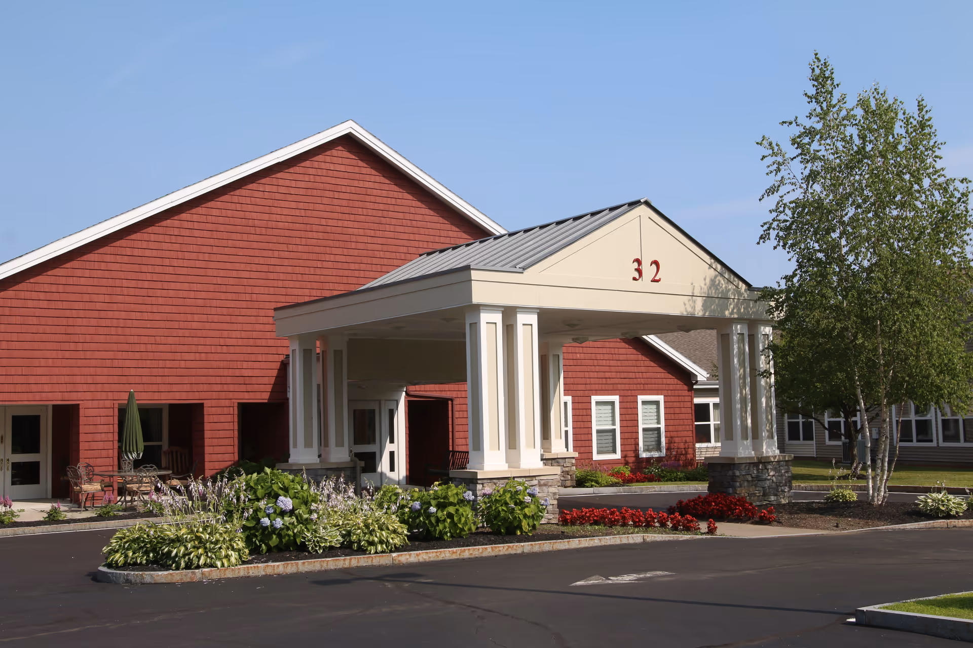 Front entrance of a red senior living building with a covered porte-cochere numbered 32 and landscaped flower beds.