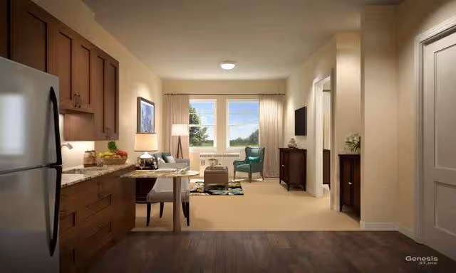 Interior view of a senior living facility apartment showing a kitchen area with wooden cabinets and a refrigerator on the left, a dining table with chairs, and a living room area with a green armchair, a small table, a TV mounted on the wall, and large windows with curtains letting in natural light.