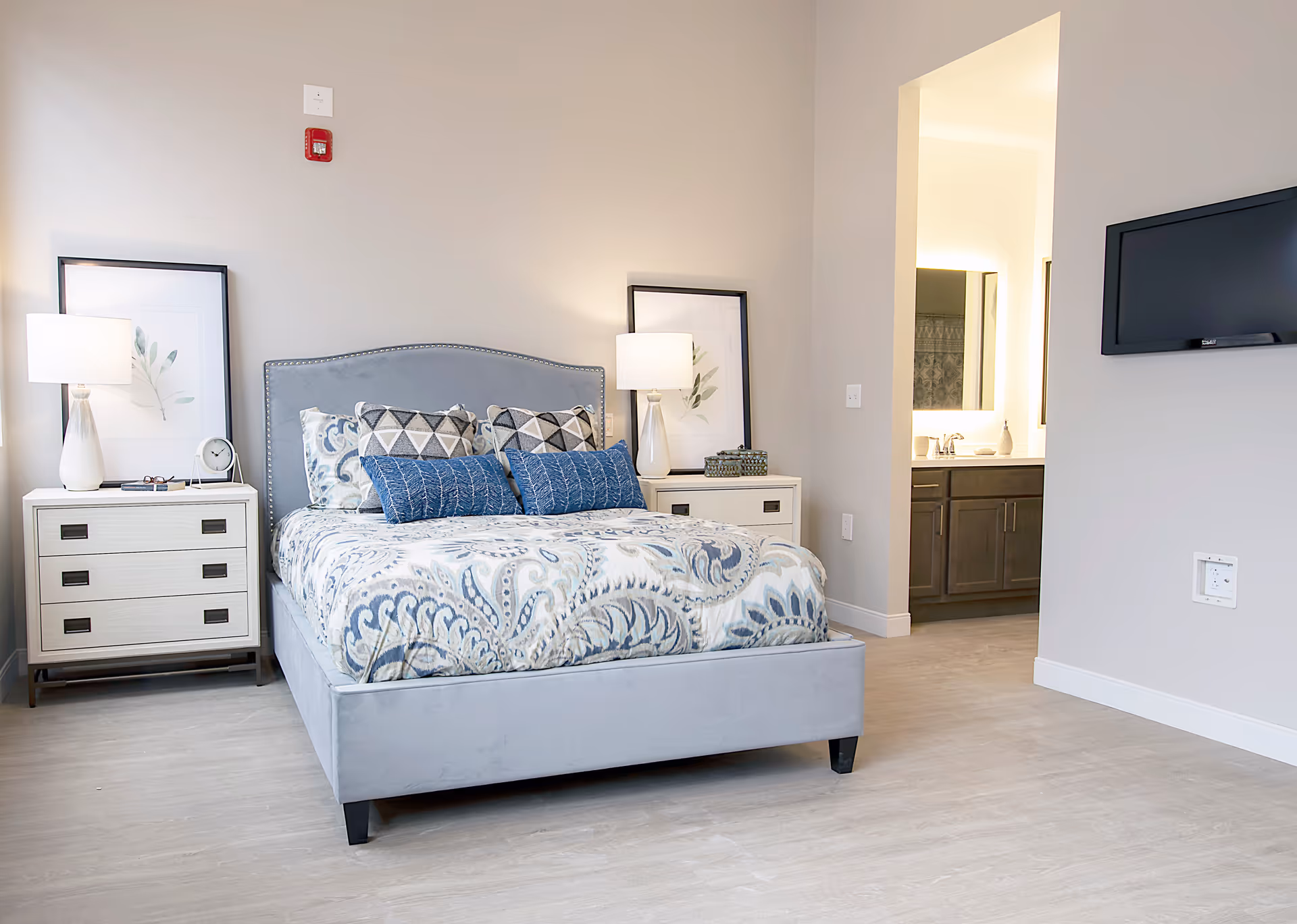 Light, modern bedroom with a gray upholstered bed, patterned bedding, matching nightstands and lamps, and an open doorway to a bathroom.