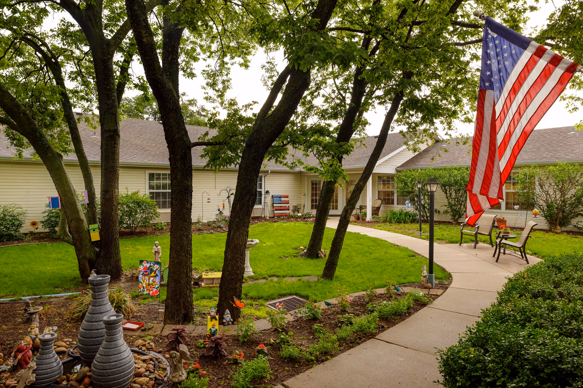 A peaceful outdoor courtyard at Homestead Assisted Living of Wellington featuring green grass, several trees, a curved concrete walkway, garden decorations, benches, and an American flag hanging on a pole.