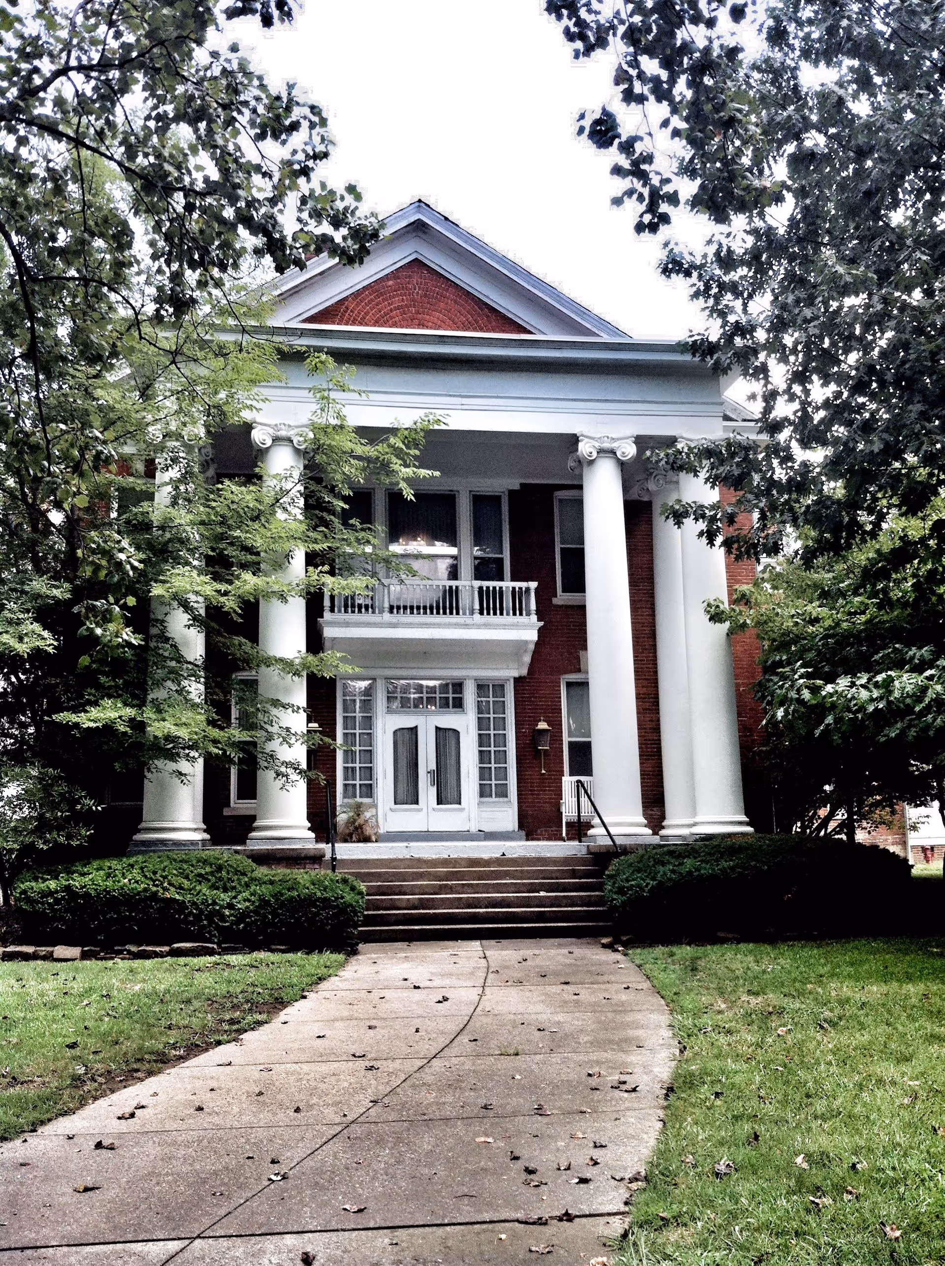 Front exterior view of a large brick building with white columns and a small balcony above the entrance. The building is surrounded by green trees and bushes, with a concrete pathway leading up to the steps of the entrance.