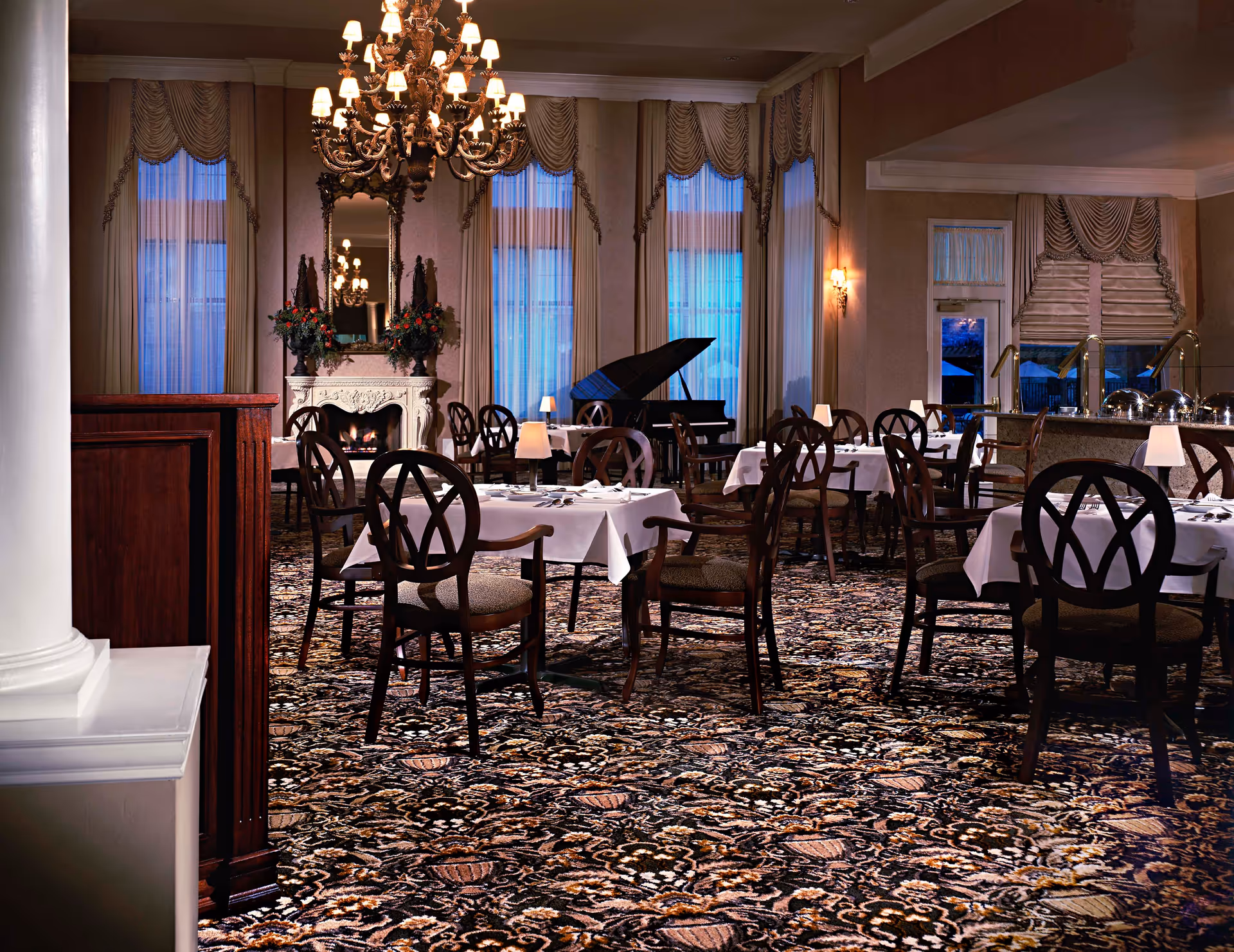 Elegant dining room with multiple tables covered in white tablecloths and set with silverware and small lamps. The room features a grand chandelier, tall windows with draped curtains, a fireplace with floral arrangements on the mantel, and a grand piano in the background. The carpet has an intricate pattern in dark tones.