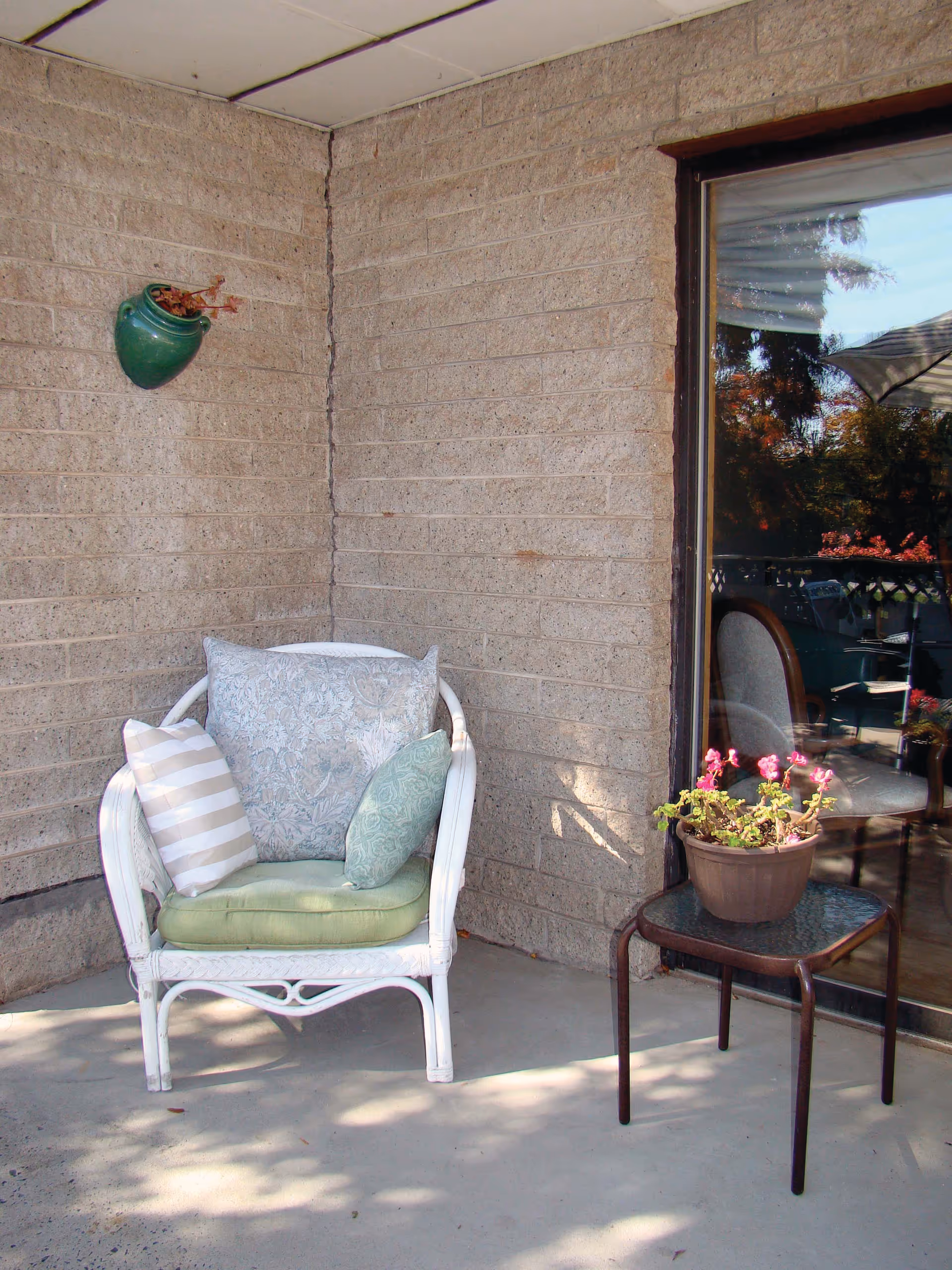 A white wicker chair with three cushions placed on a concrete patio corner next to a brick wall. A small table with a potted plant with pink flowers is beside the chair. A sliding glass door reflects outdoor furniture and trees.