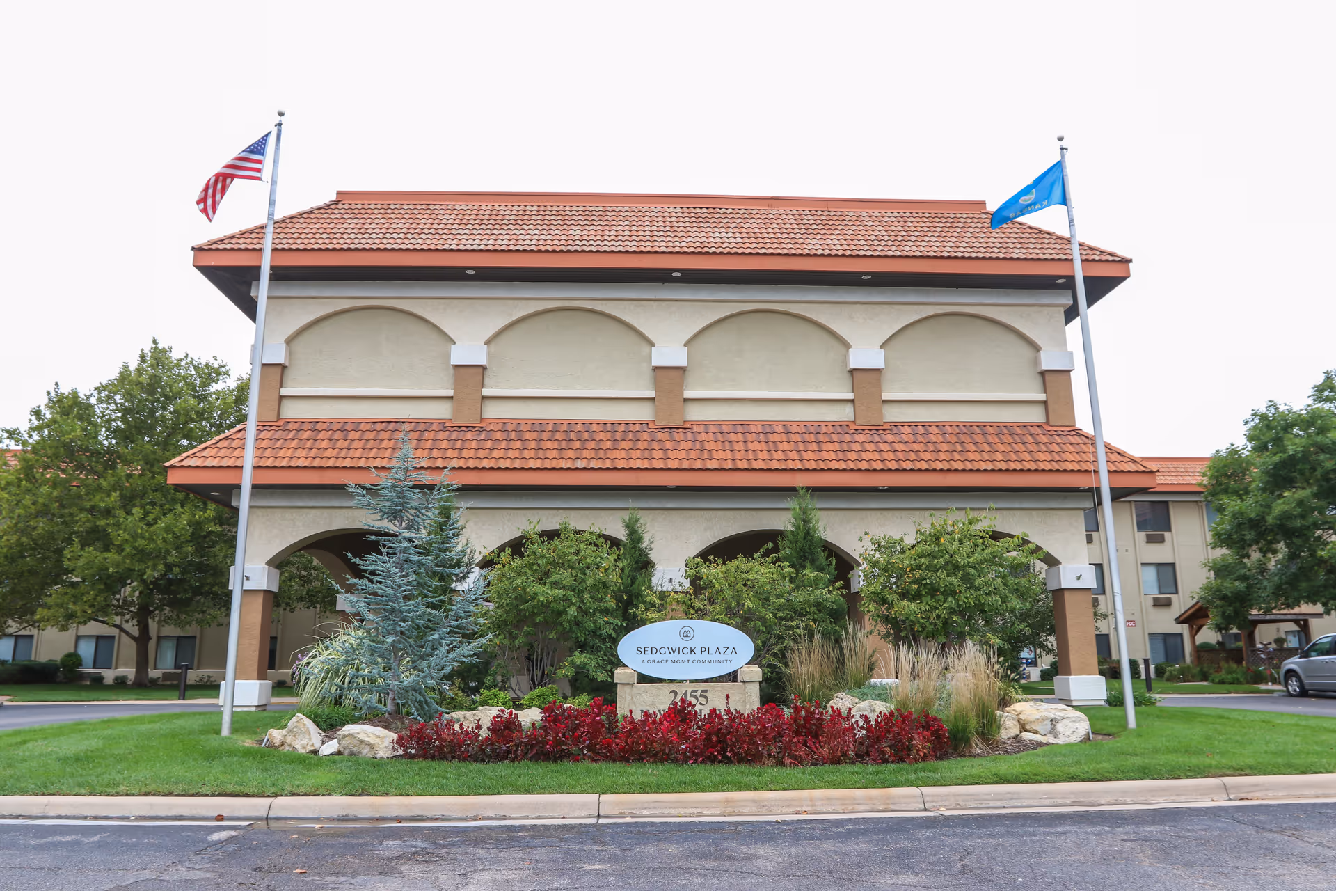 Front entrance of the Sedgwick Plaza building with two flagpoles, tiled roof, and landscaped sign at the driveway.