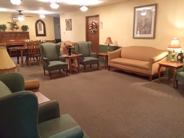 A cozy living room area in an assisted living facility featuring green upholstered armchairs, a beige sofa, wooden side tables with lamps, floral decorations, and a dining table with chairs in the background. The walls are decorated with framed artwork and a mirror.