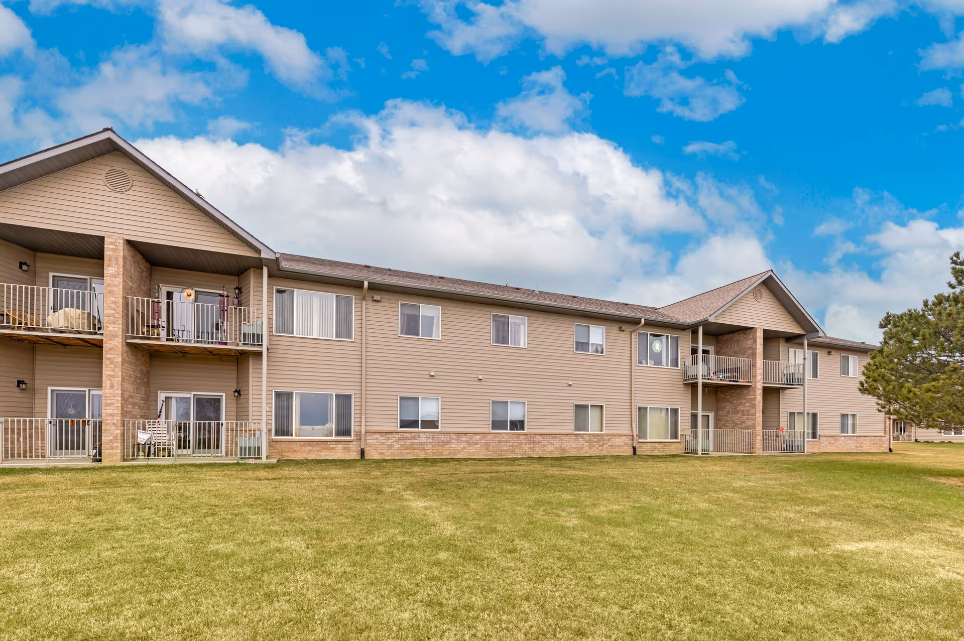Exterior view of a two-story senior living facility building with beige siding and brick accents. The building has multiple windows and balconies with railings. There is a large grassy lawn in front and a partly cloudy blue sky above.