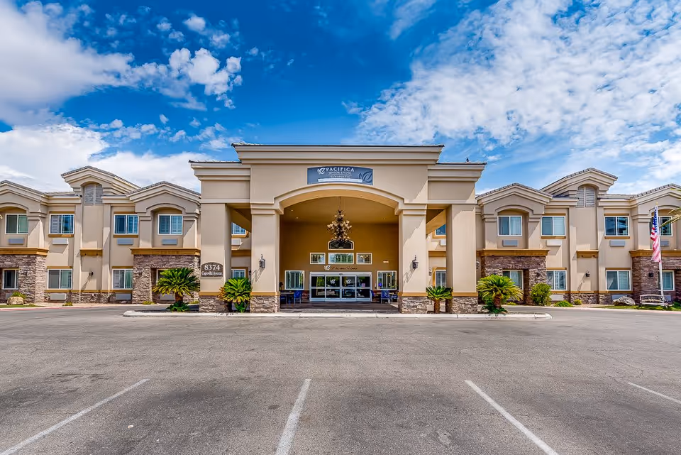 Front exterior view of a senior living facility building with a large entrance canopy, beige walls, stone accents, and multiple windows under a partly cloudy blue sky. There are small palm plants near the entrance and an American flag on the right side.
