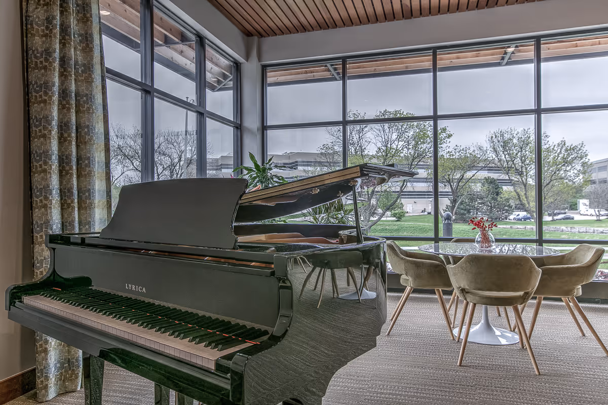Interior view of a room with a black grand piano labeled LYRICA in the foreground and a round glass table with four beige upholstered chairs near large floor-to-ceiling windows showing a green outdoor area with trees and buildings in the background.