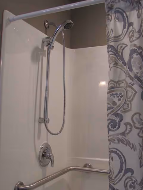 Shower stall with handheld showerhead, curved rod, patterned curtain, and grab bar.