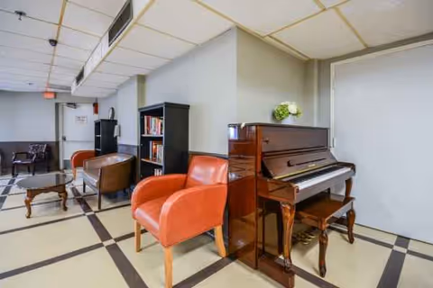 Indoor senior living lounge with a piano, red chairs, and bookshelves.