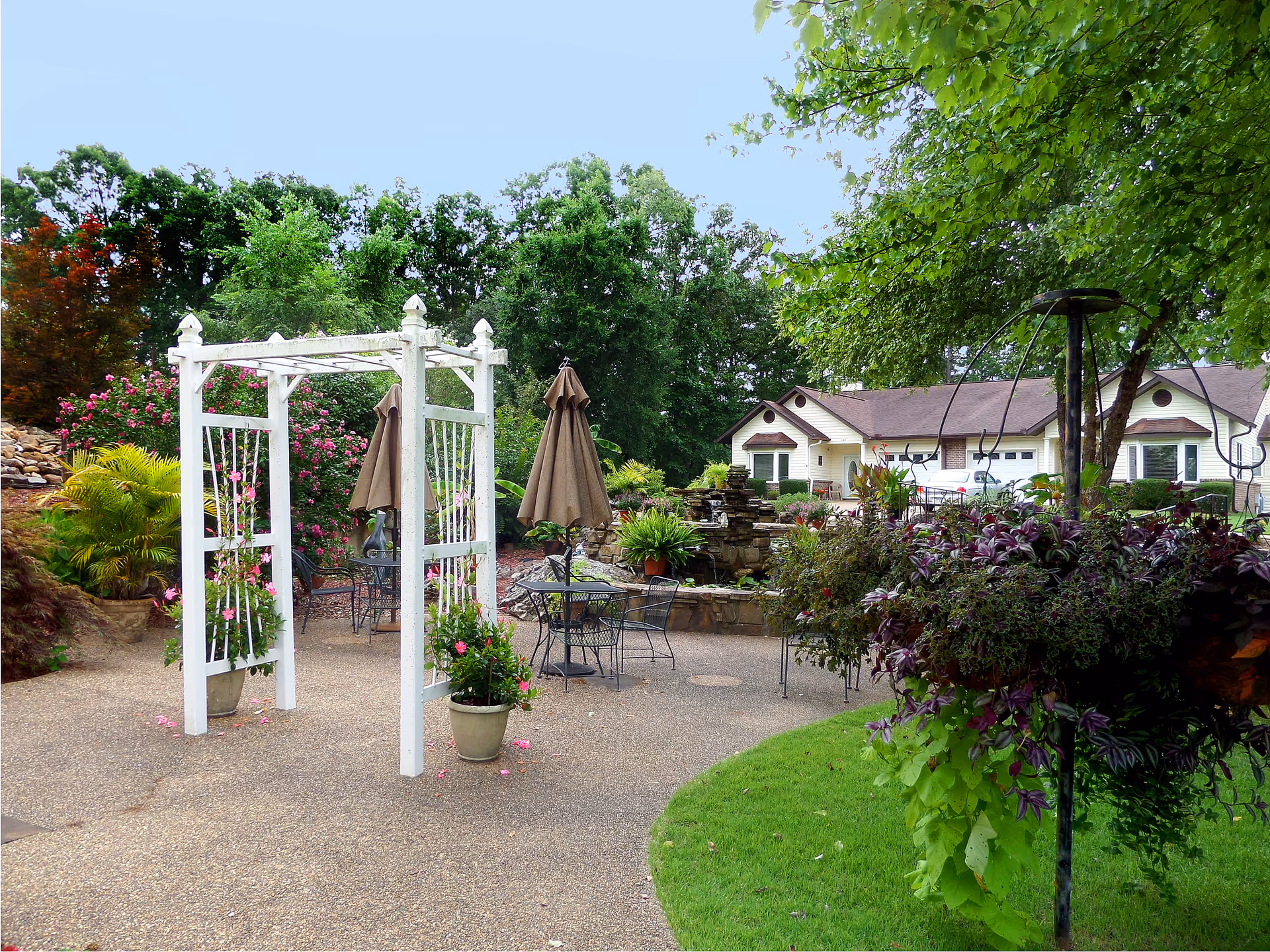 Outdoor patio area with a white wooden arbor, metal tables and chairs with brown umbrellas, surrounded by lush greenery, flowering plants, and a stone water feature. A residential building is visible in the background.