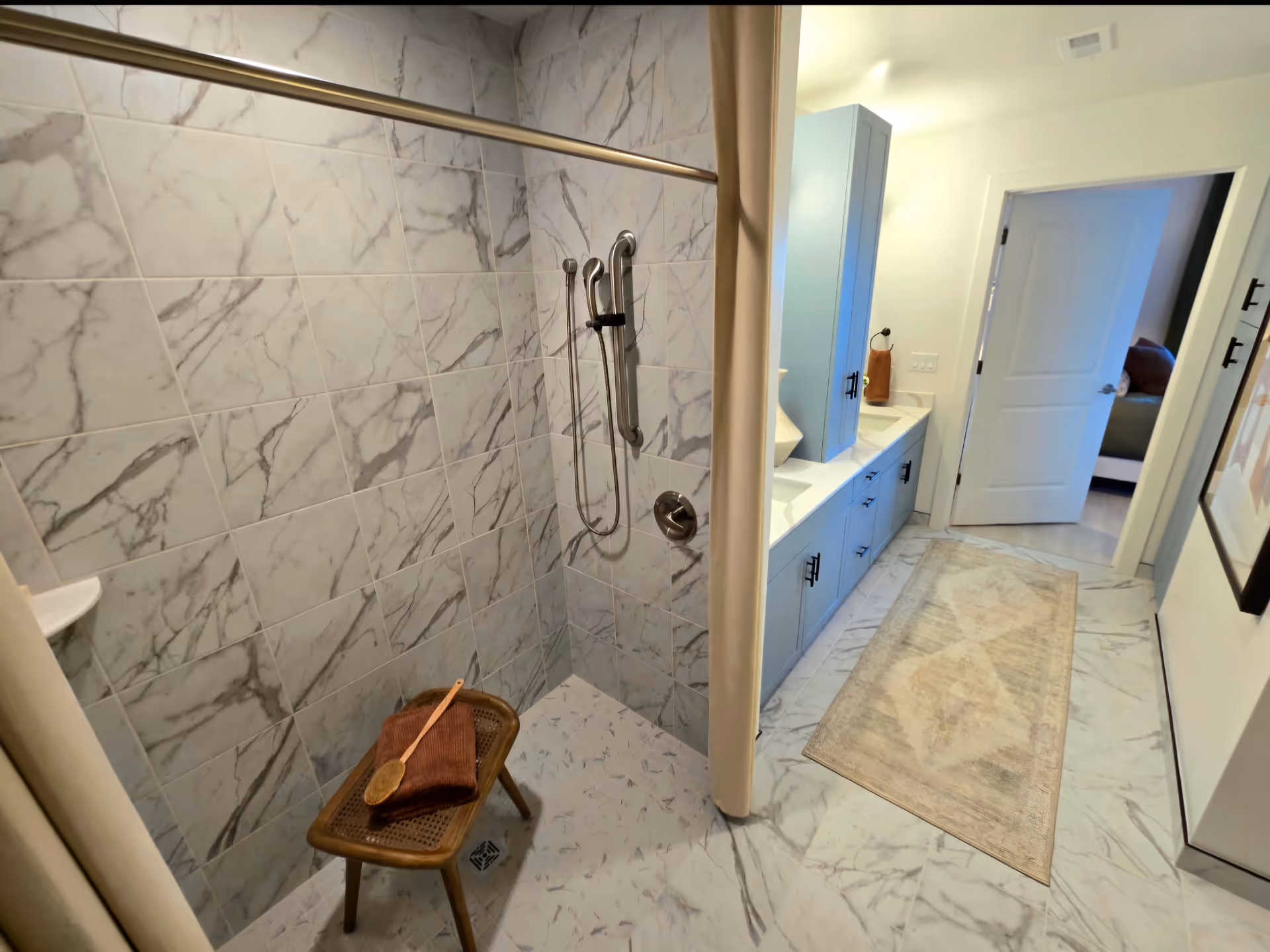 A modern bathroom featuring a walk-in shower with marble-patterned tiles, a handheld showerhead, and a small wooden stool with a brown towel and brush on it. The bathroom also has a long vanity with blue cabinets, a countertop with two sinks, and a beige patterned rug on the marble floor. A door leads to an adjacent room.