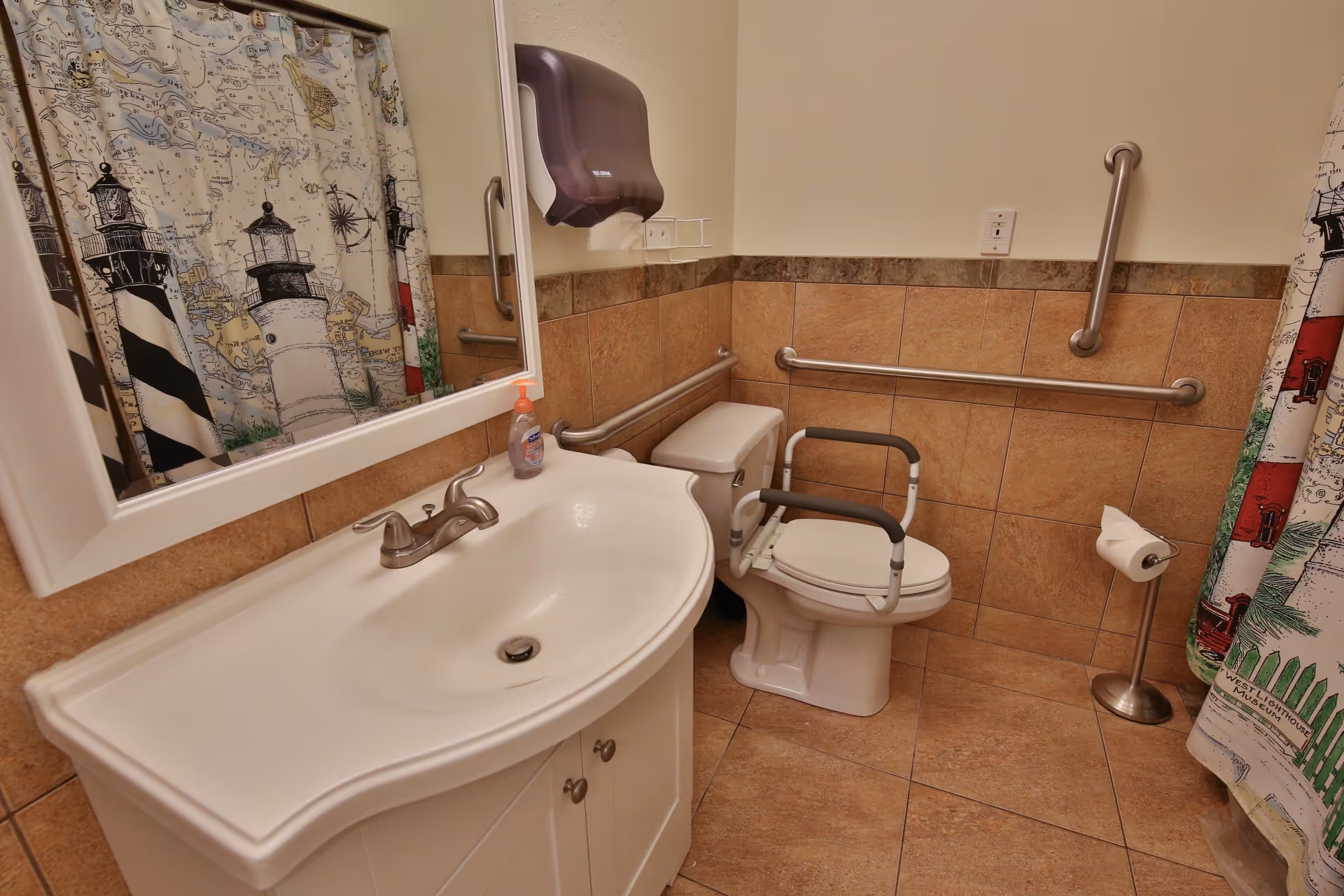 A bathroom with beige tiled walls and floor, featuring a white sink with a silver faucet, a large mirror above the sink, a toilet with safety rails, a paper towel dispenser, a toilet paper holder, and a shower curtain decorated with lighthouse images.