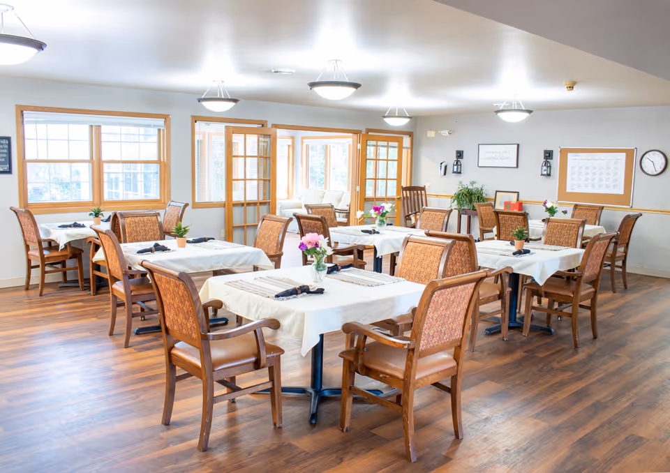 A bright assisted-living dining room with several tables set with white tablecloths, flowers, and wooden chairs.