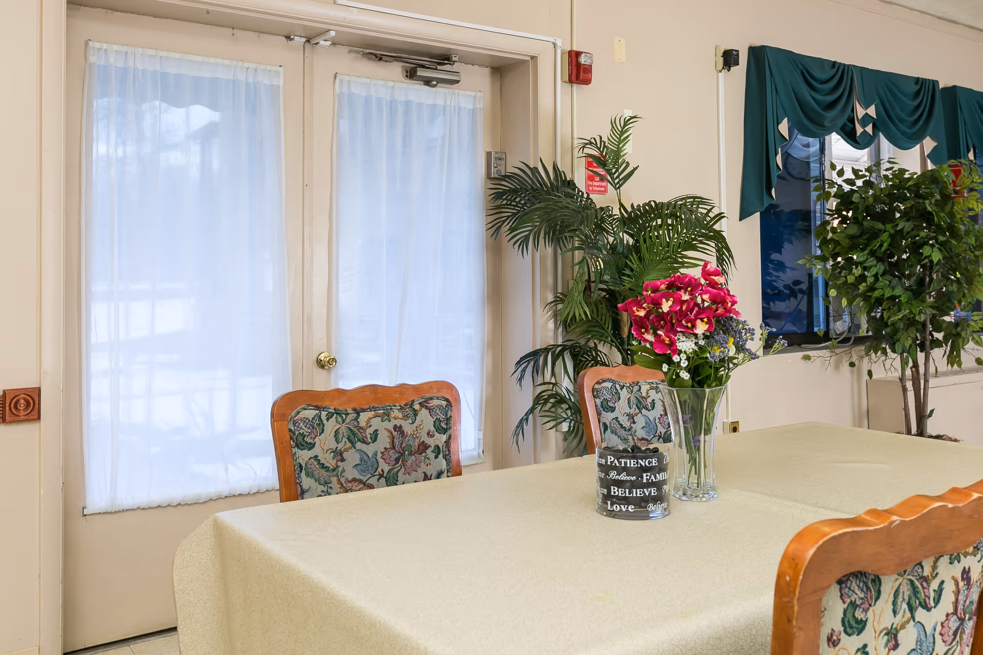 A dining table with a floral centerpiece and upholstered chairs in a communal room with glass doors and potted plants.