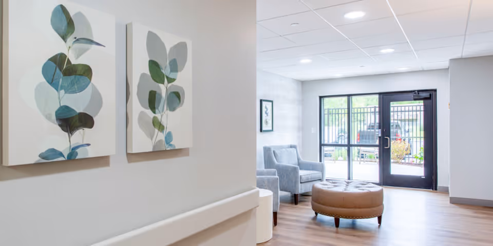 A bright and modern common area with light wood flooring, two gray upholstered armchairs, a round brown ottoman, and large windows with a glass door leading outside. Two botanical-themed paintings hang on a white wall with a handrail.