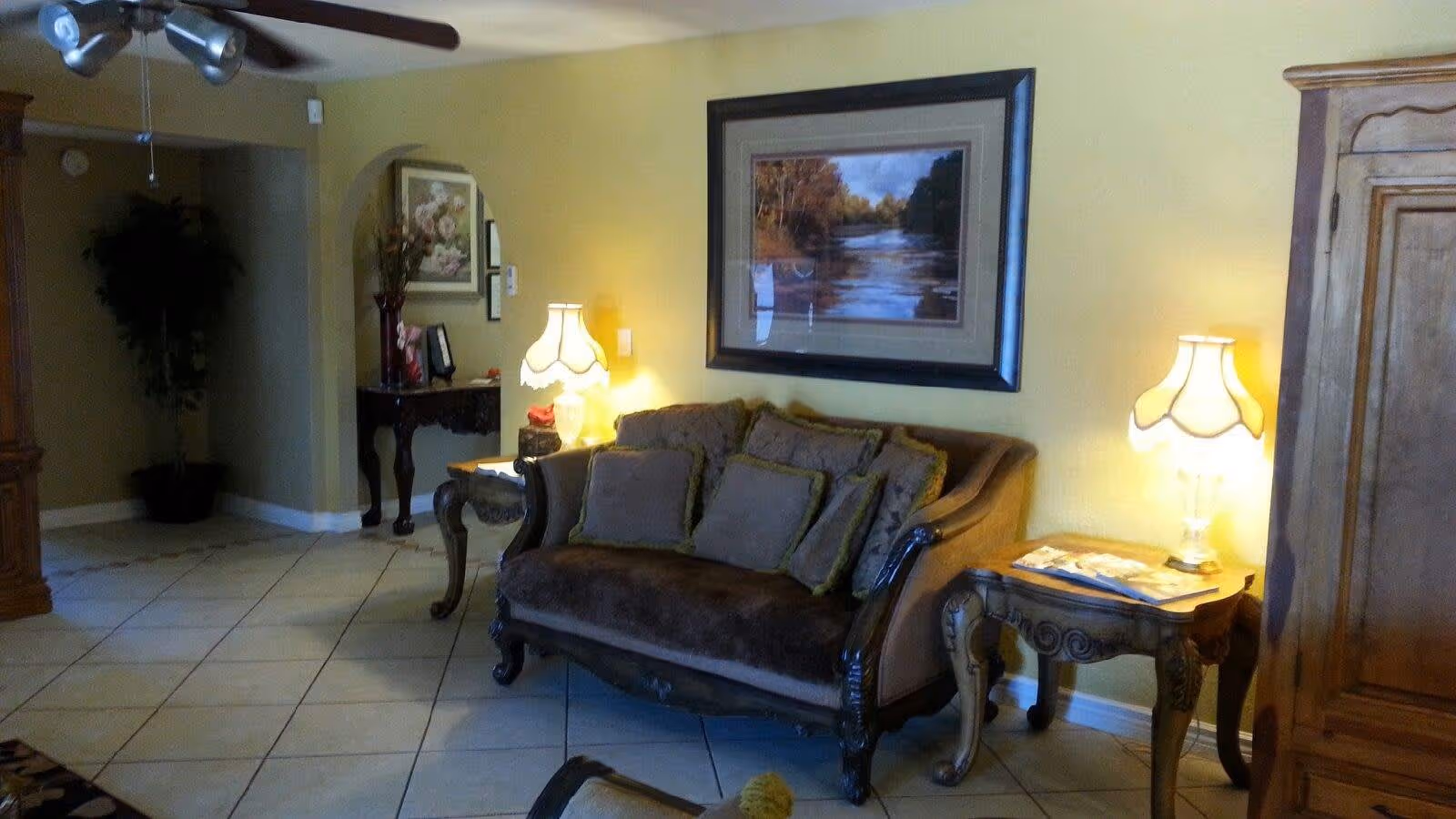 A furnished living room with a vintage sofa, ornate side tables and lamps, a large framed painting on a yellow wall, and a tiled floor.