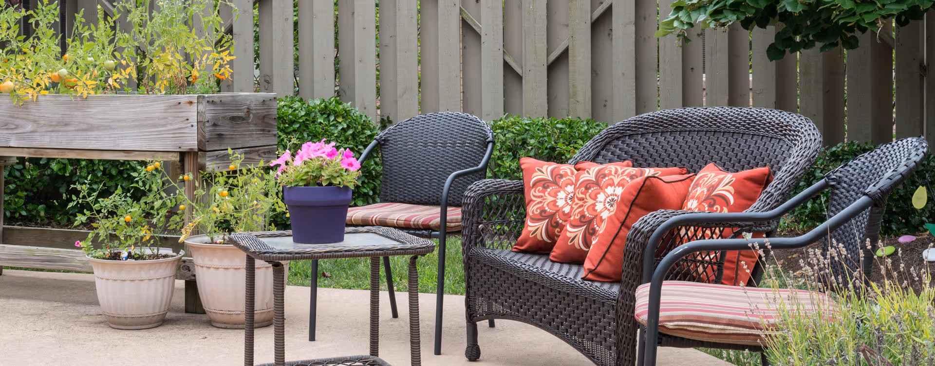 Outdoor patio area with dark wicker furniture including a loveseat and two chairs with red and patterned cushions. A small wicker table holds a purple pot with pink flowers. Several potted plants and a wooden planter box with greenery are visible against a wooden fence in the background.