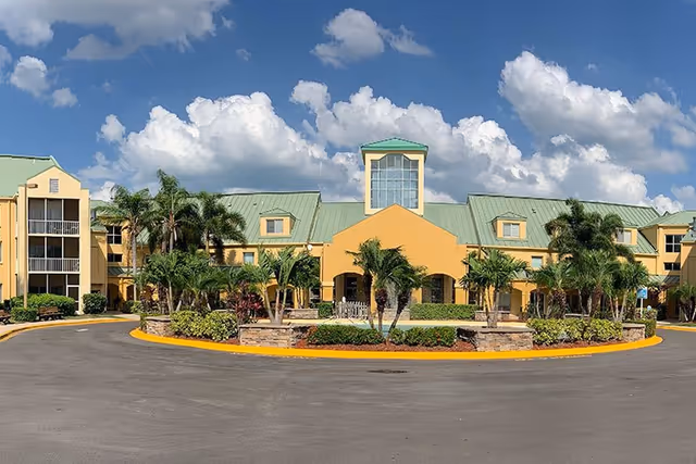 Front exterior of a yellow senior living building with green roofs, palm trees, and a circular driveway under a blue sky.