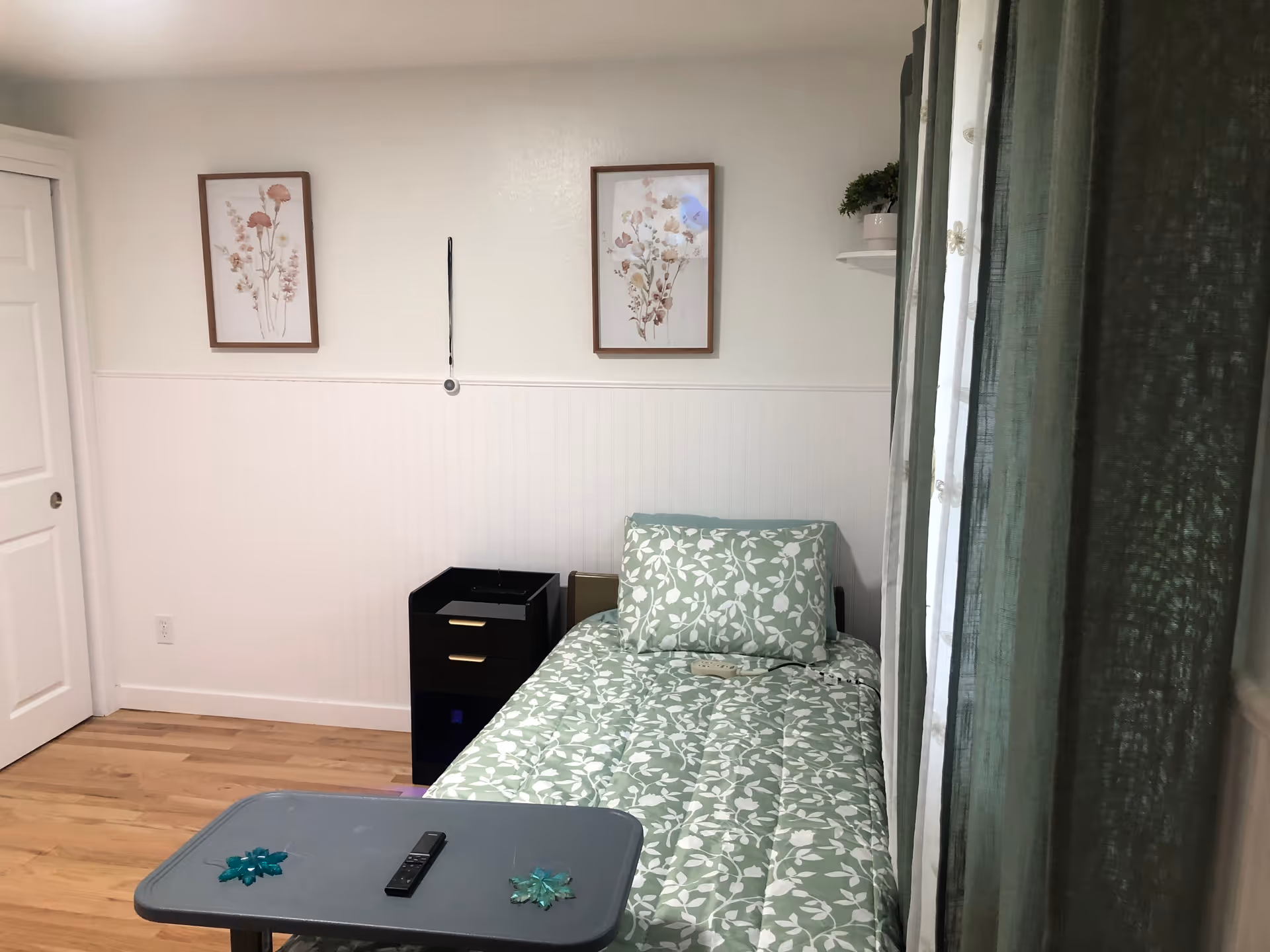 A small bedroom with a single bed covered in a green and white floral patterned comforter and matching pillow. Next to the bed is a black nightstand with two drawers and a small shelf. Above the bed on the wall are two framed floral prints. A gray tray table with a remote control and two decorative items is positioned in front of the bed. Green curtains hang on the right side of the room, partially covering a window.