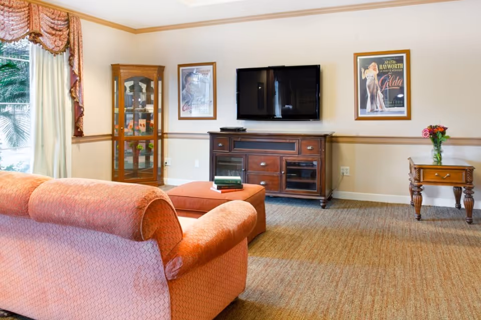 A cozy living room with a peach-colored sofa and matching ottoman facing a wooden TV stand with a flat-screen television mounted on the wall above it. The room features a glass display cabinet in the corner, two framed vintage movie posters on the wall, a small wooden side table with a vase of flowers, and large windows with decorative curtains letting in natural light.
