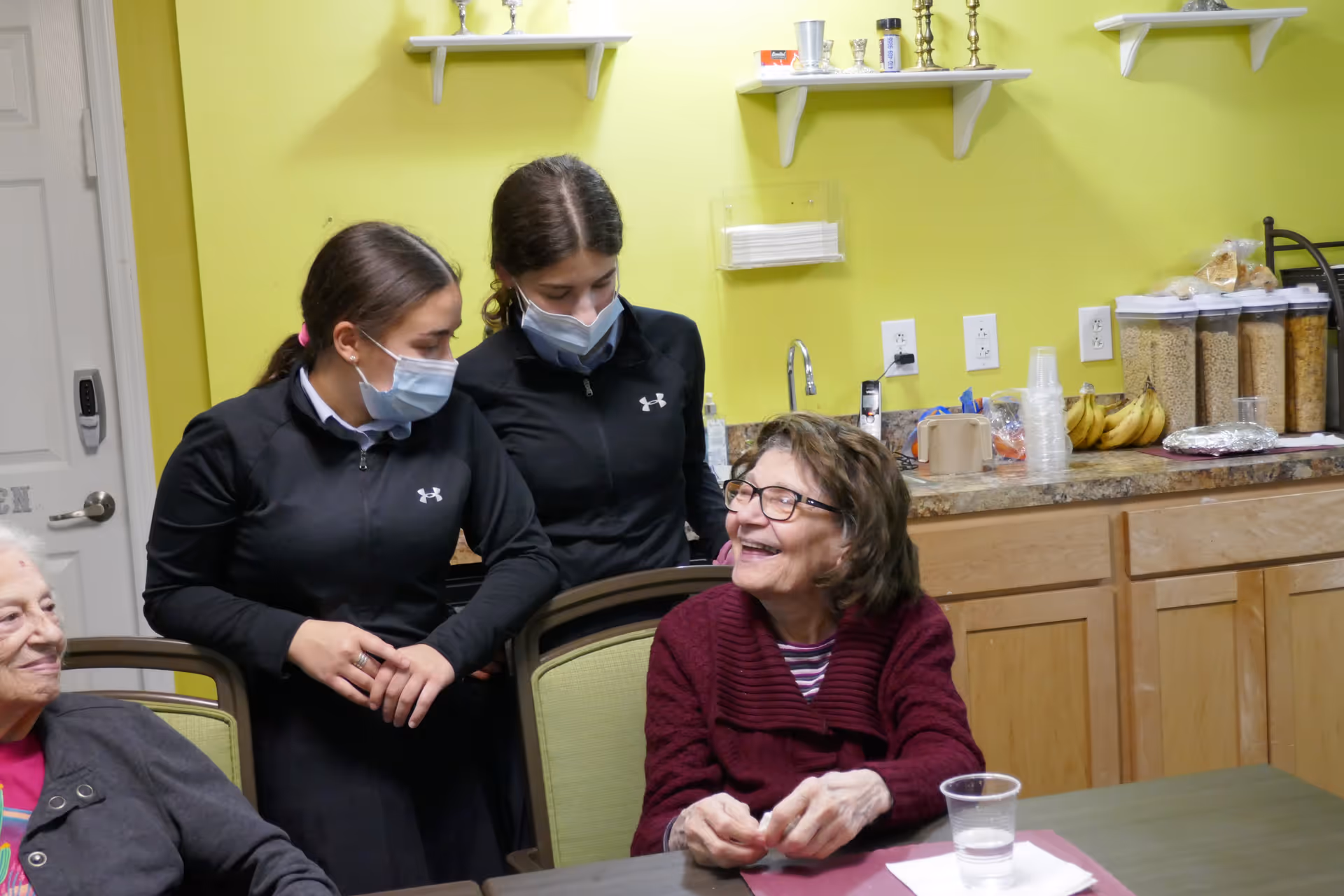 Two young women wearing black jackets and face masks stand behind two elderly women seated at a table in a room with yellow walls and wooden cabinets. The elderly woman on the right is smiling and wearing glasses and a maroon sweater. The countertop behind them has containers of cereal, bananas, plastic cups, and other items.