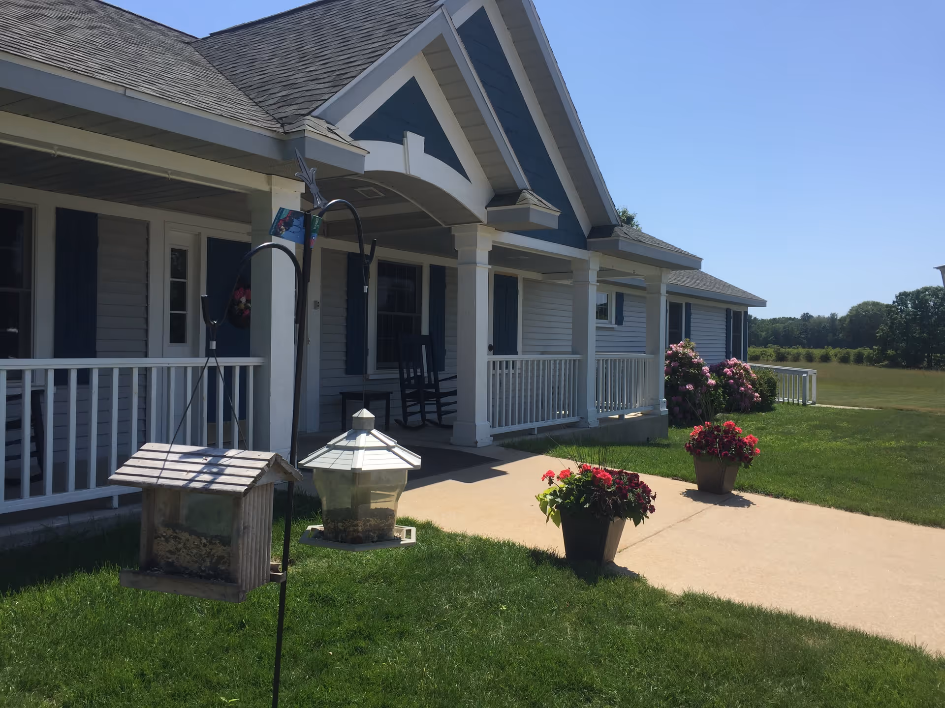Front exterior view of a single-story building with a covered porch, white railings, and blue shutters. There are two bird feeders hanging on a metal pole in the foreground, and two flower pots with red and pink flowers on the concrete walkway leading to the porch. The building is surrounded by green grass and some bushes, with a clear blue sky in the background.