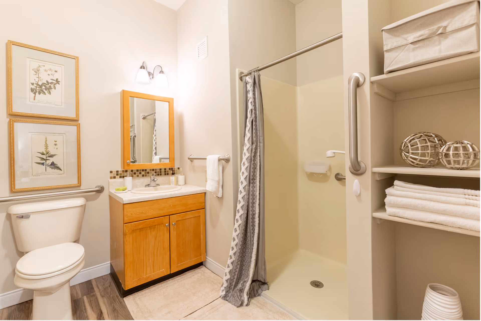 A clean and well-lit bathroom featuring a toilet, a wooden vanity with a sink and mirror above it, a shower area with a gray patterned curtain, and built-in shelves holding folded white towels and decorative items.