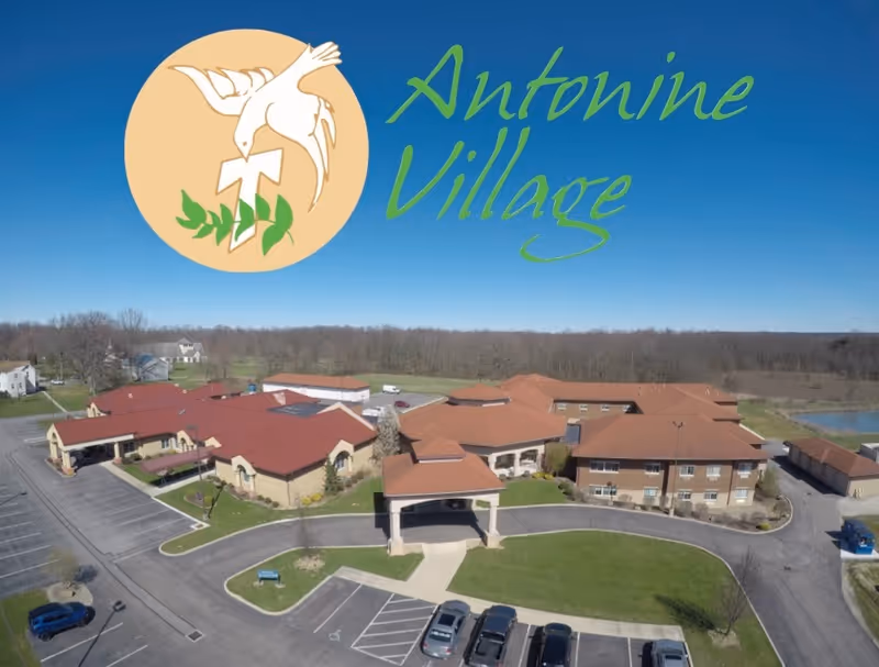Aerial view of Antonine Village, a senior living facility with multiple connected buildings featuring red roofs and beige walls, surrounded by parking lots, green lawns, and trees under a clear blue sky.