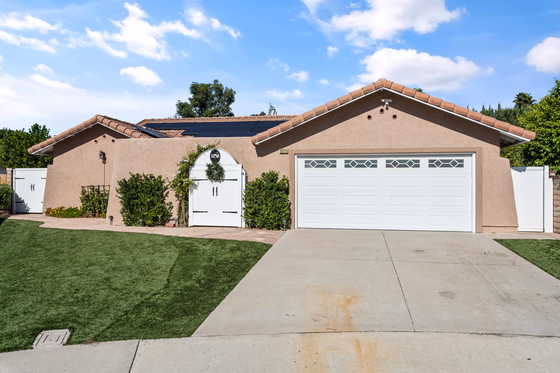 Front exterior view of a single-story house with a beige stucco finish, a white double garage door, a white arched gate with a wreath, green bushes, and a well-maintained lawn under a blue sky with scattered clouds.