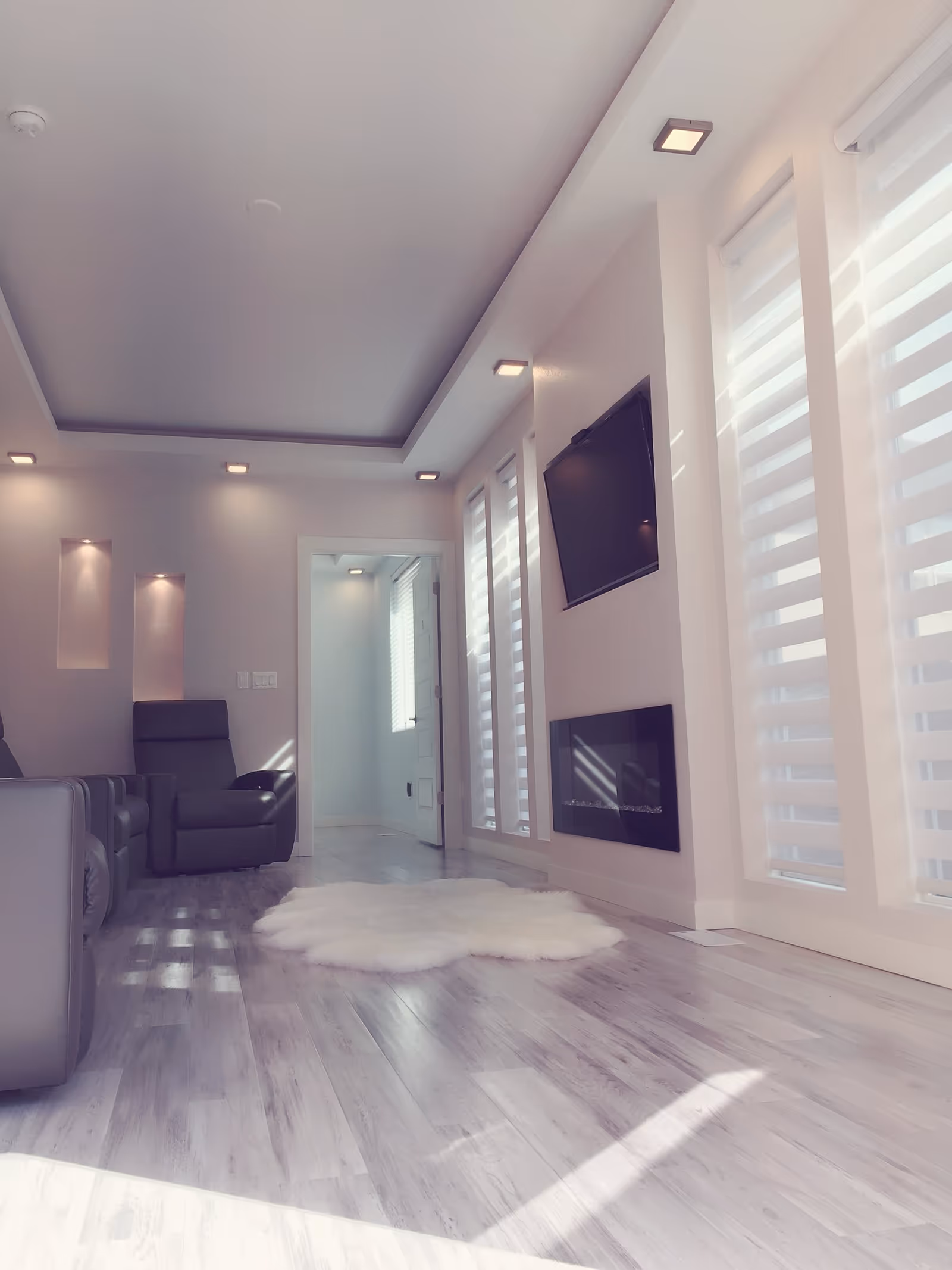 A modern living room with light wood flooring, a white fluffy rug in the center, gray recliner chairs along the left wall, a wall-mounted flat screen TV above a sleek black fireplace on the right wall, and large windows with white blinds letting in natural light.