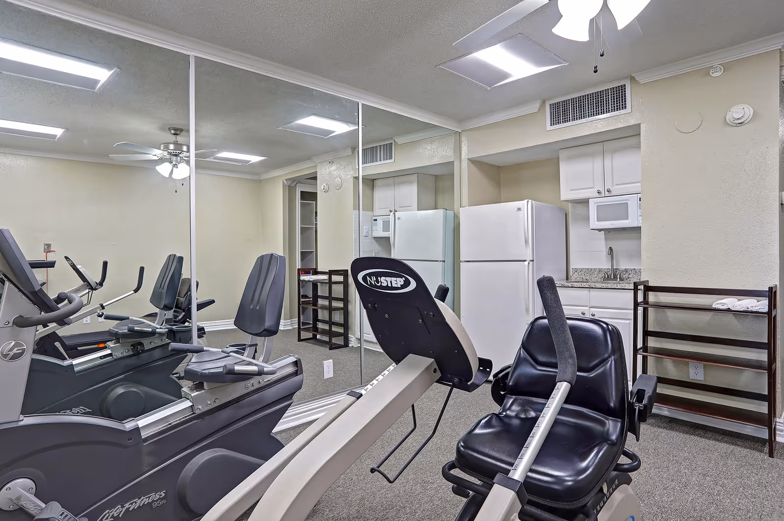 A small fitness room with exercise equipment including a recumbent bike and a treadmill. The room has mirrored walls, a ceiling fan, fluorescent lighting, and a small kitchenette area with a refrigerator, microwave, sink, and cabinets.