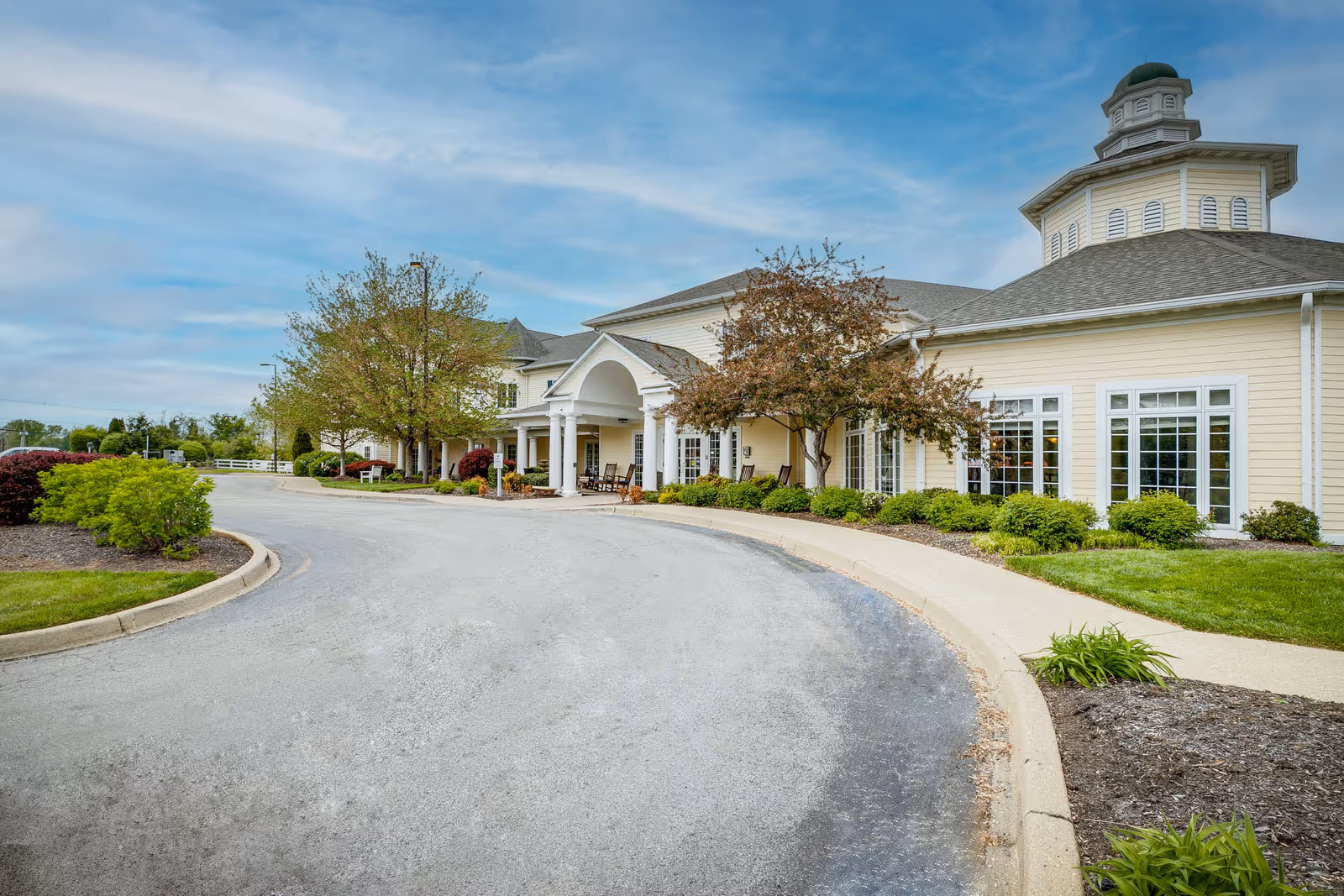 Exterior view of a senior living facility with a curved driveway, landscaped bushes, trees, and a beige building featuring large windows and a cupola on the roof under a partly cloudy sky.