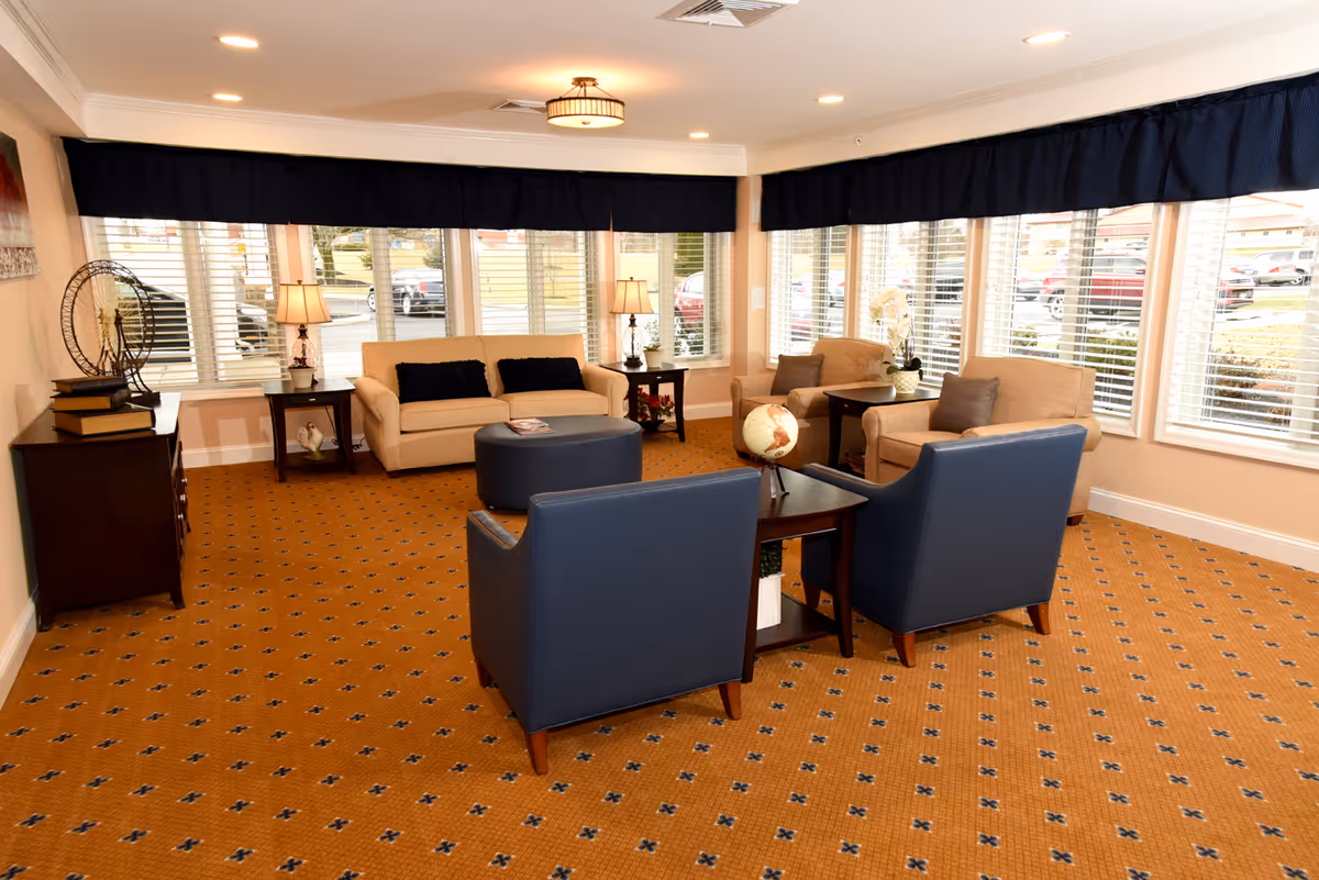 A bright and spacious senior living facility common area with large windows covered by dark blue valances. The room features a beige sofa with two dark cushions, two beige armchairs with gray cushions, and two dark blue armchairs arranged around a dark wooden coffee table with a globe on it. There are two side tables with lamps and decorative items, and a patterned orange carpet covers the floor.