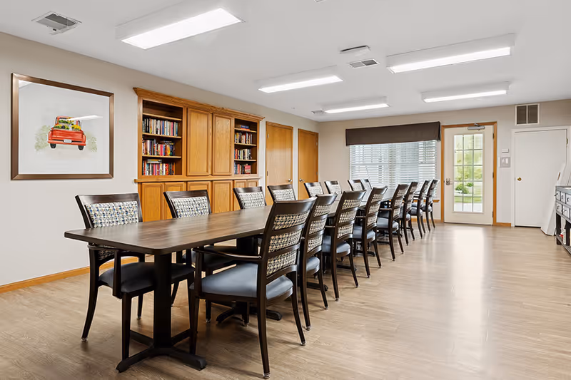 A long rectangular dining table with sixteen chairs arranged around it in a well-lit room. The room has light-colored walls and wood flooring. On the left wall, there is a framed picture of a red truck carrying flowers. Behind the table, there is a wooden built-in bookshelf filled with books and cabinets. A window with blinds and a door leading outside are visible at the far end of the room.