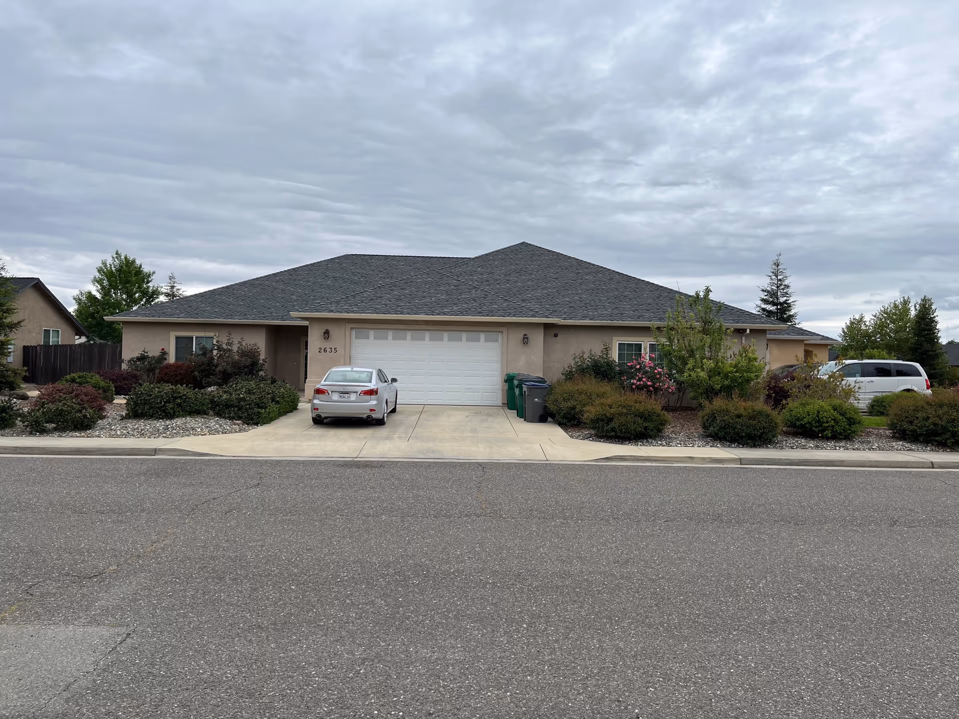 Front exterior view of a single-story residential building with a gray shingled roof, beige walls, a double garage door, and two cars parked in the driveway. The house is surrounded by landscaped bushes and trees under a cloudy sky.