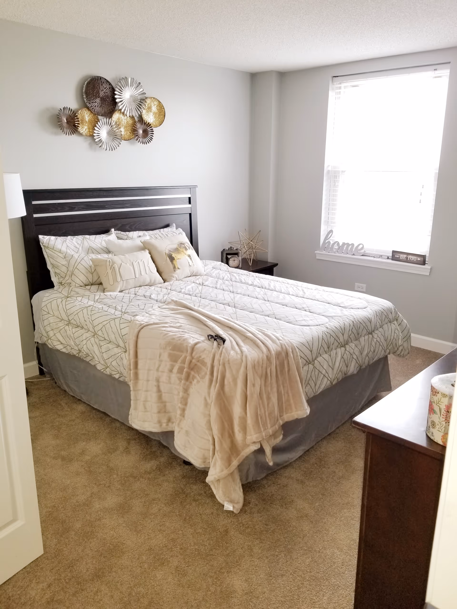 A neatly made bed with patterned white and gray bedding and decorative pillows in a bedroom. The bed has a dark wooden headboard and a beige throw blanket draped over the foot. There is a nightstand with a clock and decorative star next to the bed. A window with white blinds lets in natural light, and the word 'home' is displayed on the windowsill along with a small sign that says 'BE YOU'. The walls are light gray and the floor is carpeted in beige.