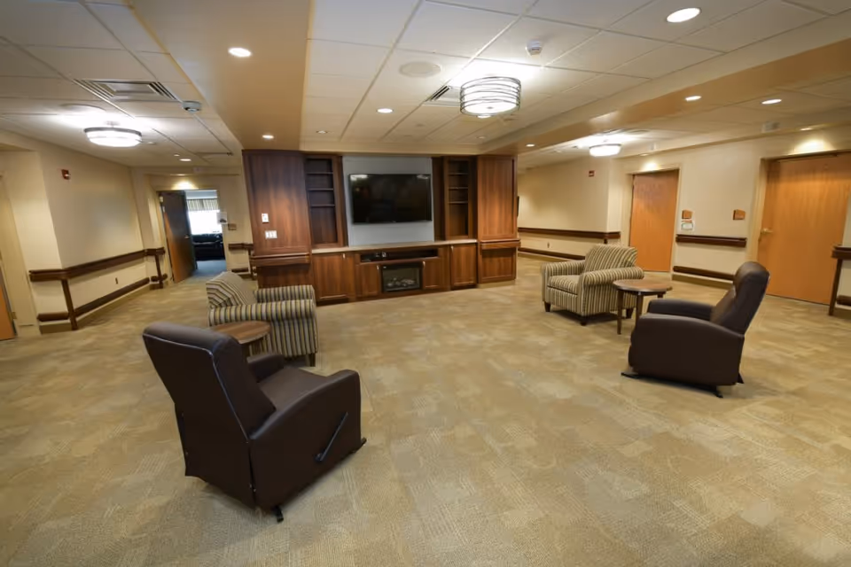 Spacious senior living common lounge with armchairs, small tables, and a wall-mounted TV in wood cabinetry.