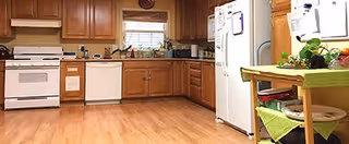 A spacious kitchen with wooden cabinets, a white stove, dishwasher, and refrigerator. There is a window above the sink with a small plant on the windowsill. A small table with a green tablecloth and some dishes is visible on the right side.