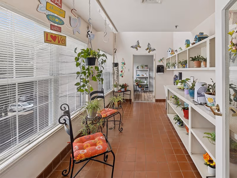 A bright indoor hallway with large windows on the left side covered by white blinds. Two black metal chairs with orange floral cushions are placed along the window wall. Various potted plants are arranged on the chairs, hanging from the ceiling, and on white shelves on the right side. The floor is covered with reddish-brown tiles. The far end of the hallway leads to another room with more plants and wooden flooring. Decorative butterfly wall hangings and a colorful sign that reads 'Let things grow with love' are visible.