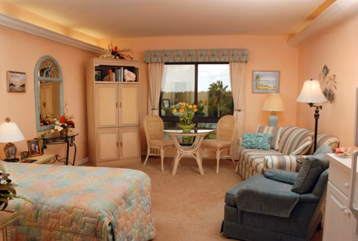Studio apartment-style bedroom with a bed in the foreground, a striped sofa and armchair, a small table and chairs by a window, and light wood cabinetry.
