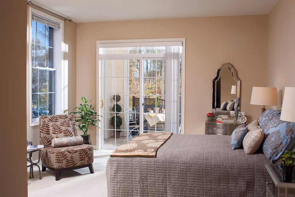 A cozy bedroom with a large bed covered in a gray quilt and decorative pillows. There is a patterned armchair with a bolster pillow near a window on the left. A small table with a plant is beside the chair. Sliding glass doors lead to an outdoor patio with a table and chairs. A dresser with a decorative mirror, lamp, and books is on the right side of the room. The walls are painted beige and the room is well-lit with natural light.