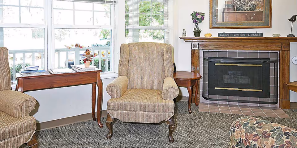 Cozy seating area with upholstered armchairs, side tables, large windows, and a fireplace mantel.