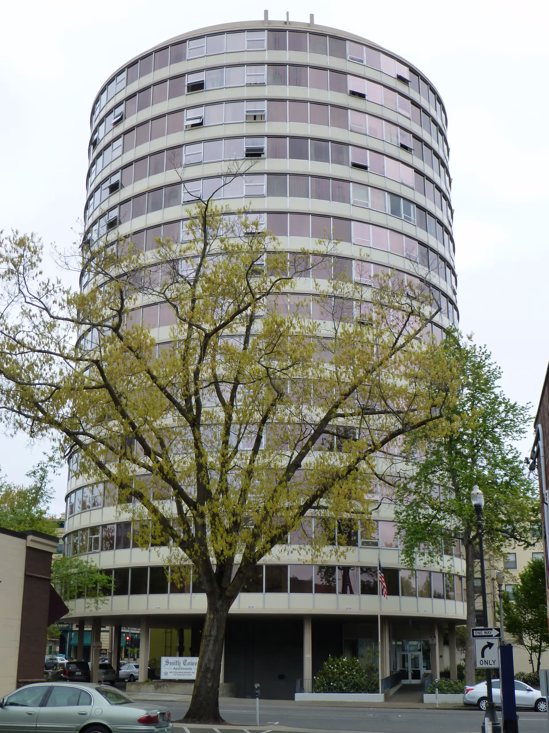 Round multi-story apartment building with many windows and a large tree in front.