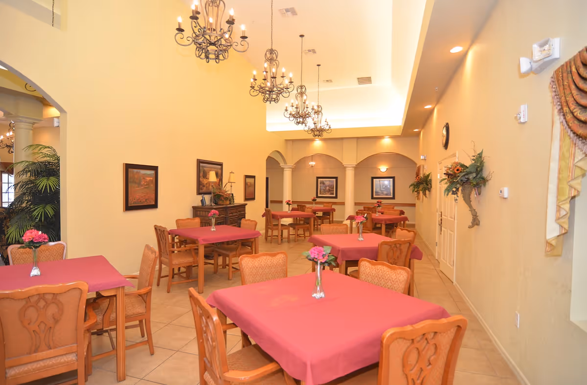 A dining room with multiple square tables covered with red tablecloths, each table having a small vase with pink flowers. The room features beige walls, tiled floors, decorative wall art, and several ornate chandeliers hanging from a high ceiling. There are wooden chairs with cushioned seats around the tables, and some greenery is visible near an archway on the left side.