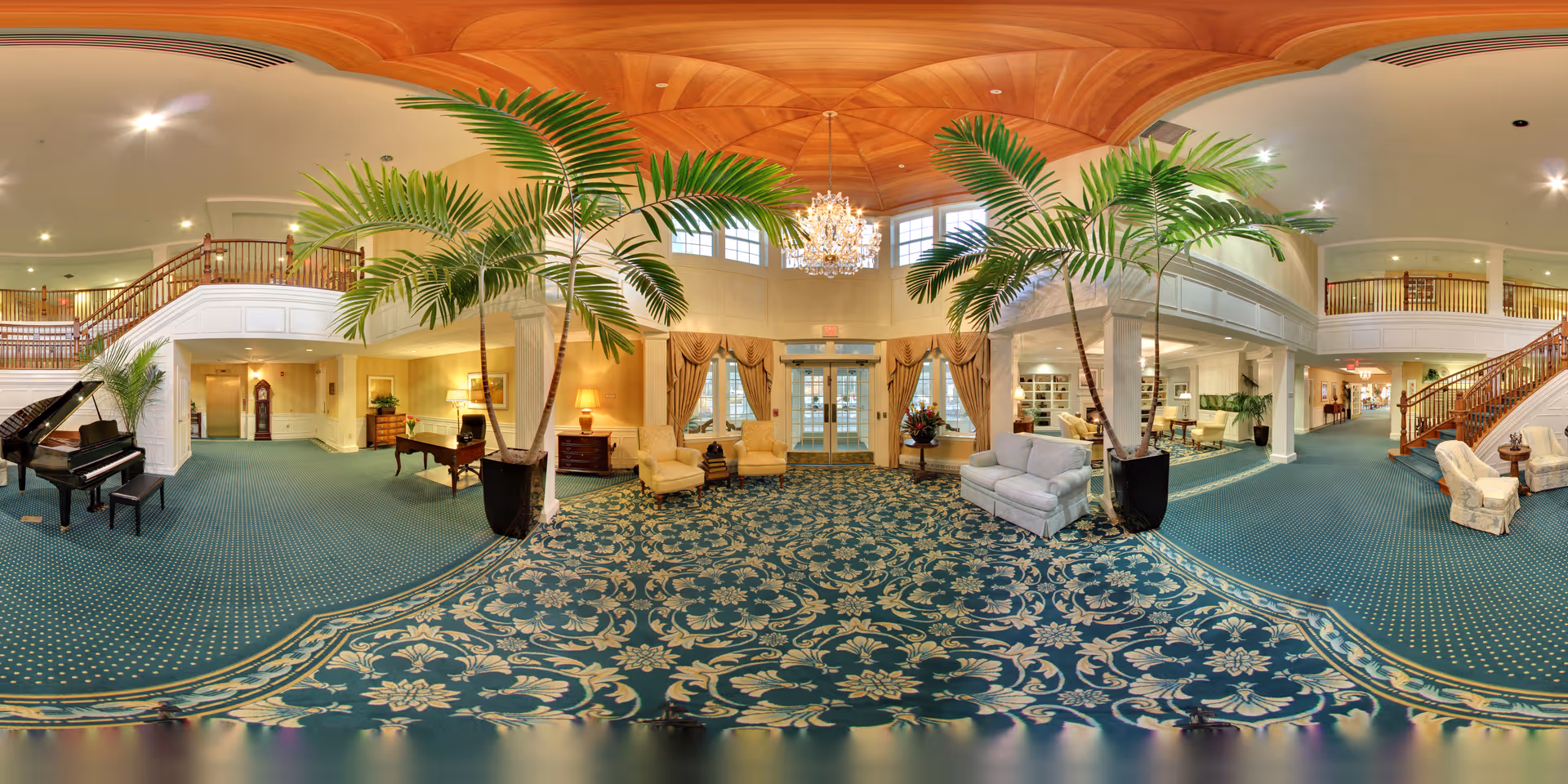 Spacious and elegant senior living facility lobby with a high wooden ceiling, large chandelier, patterned blue carpet, tall indoor palm plants, grand piano, comfortable seating areas with armchairs and sofas, and a staircase leading to an upper level balcony.