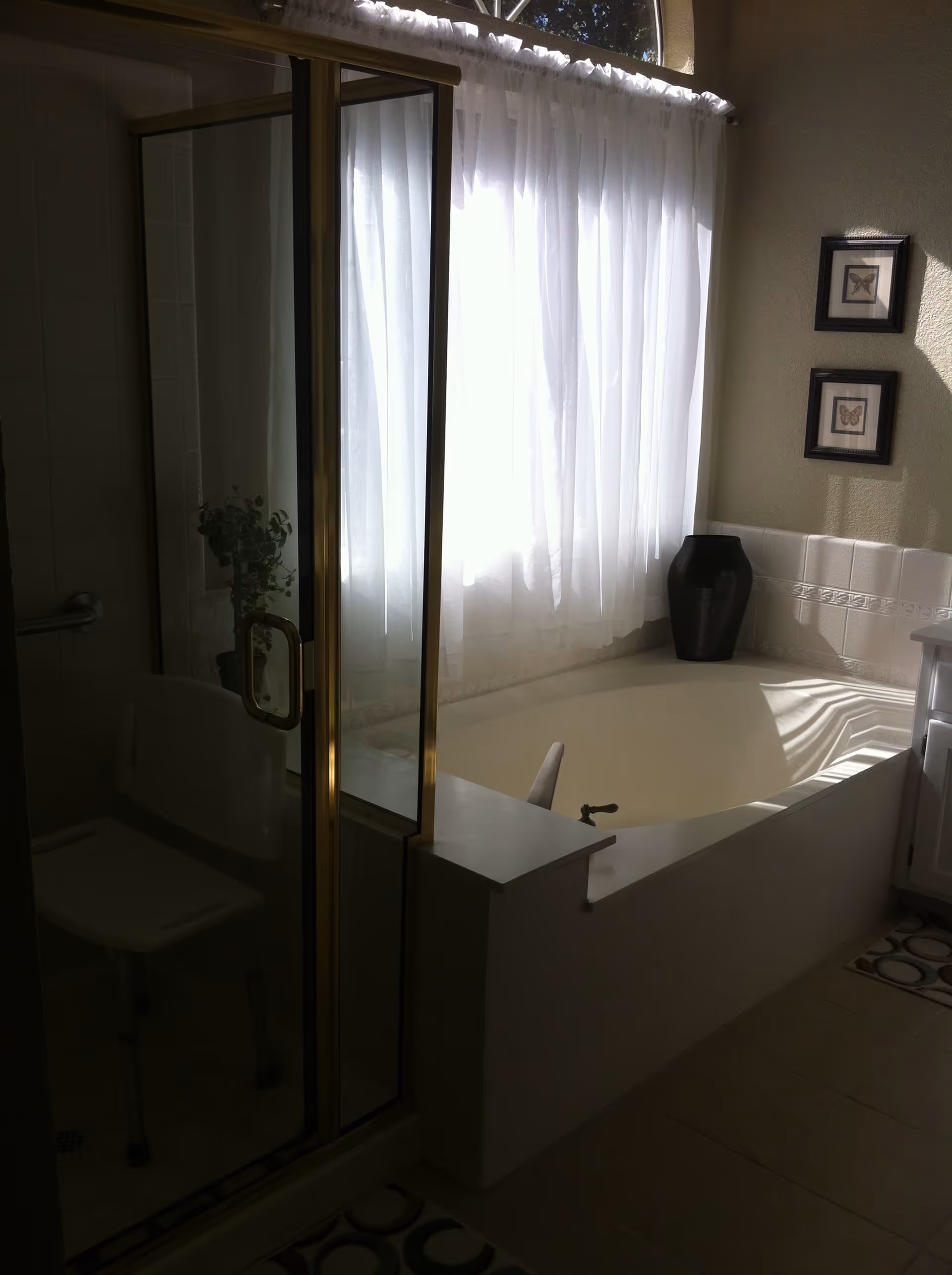 A bathroom with a glass-enclosed shower featuring a white shower chair inside, a large bathtub next to a window with sheer white curtains, a black vase on the bathtub ledge, and two framed butterfly pictures on the wall.