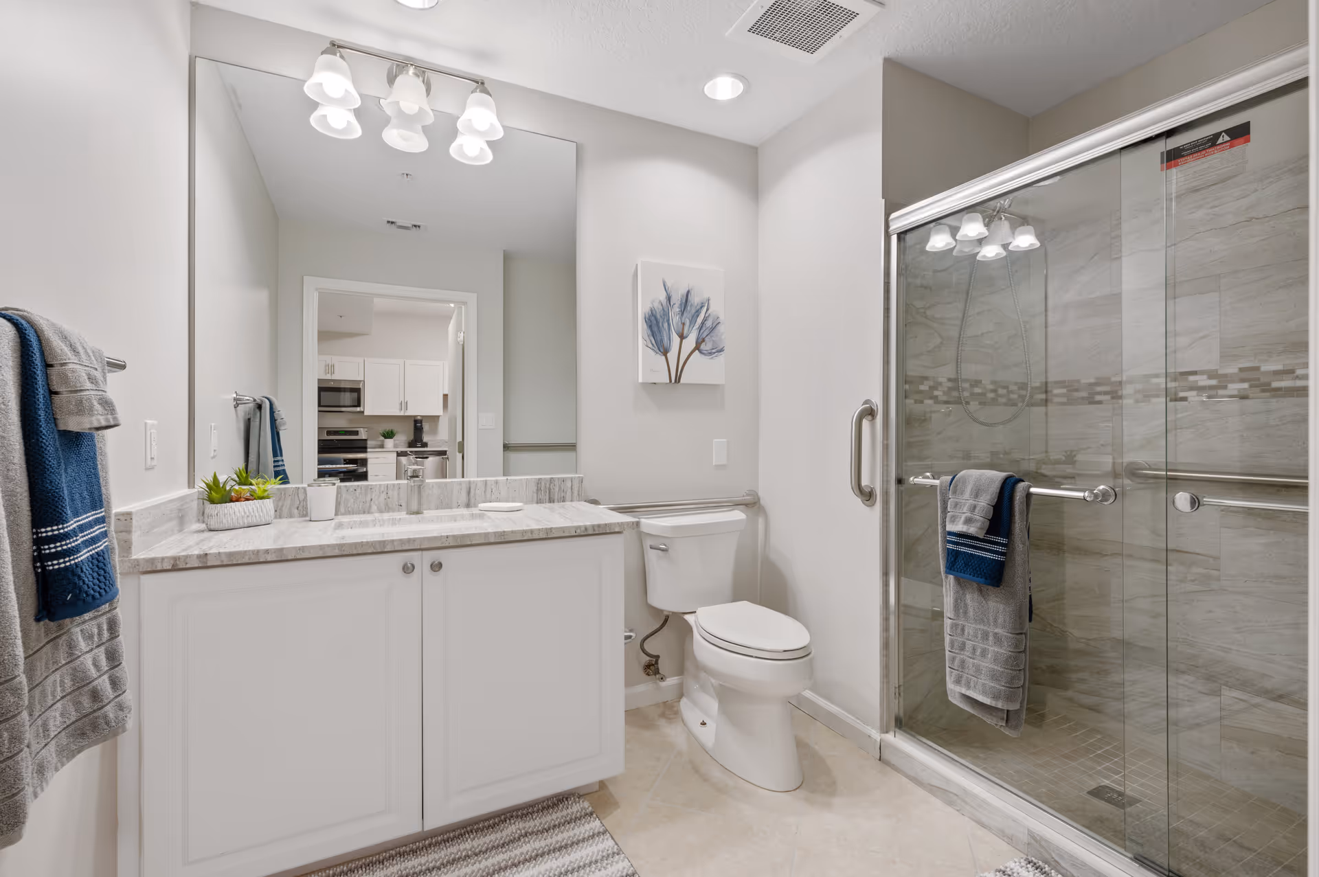A modern bathroom featuring a white vanity with a marble countertop, a large mirror with overhead lighting, a toilet, and a glass-enclosed shower with tiled walls. Towels hang on racks near the vanity and shower. A piece of wall art with blue floral design is mounted above the toilet. The bathroom floor is tiled, and the adjacent room visible through the doorway appears to be a kitchen.